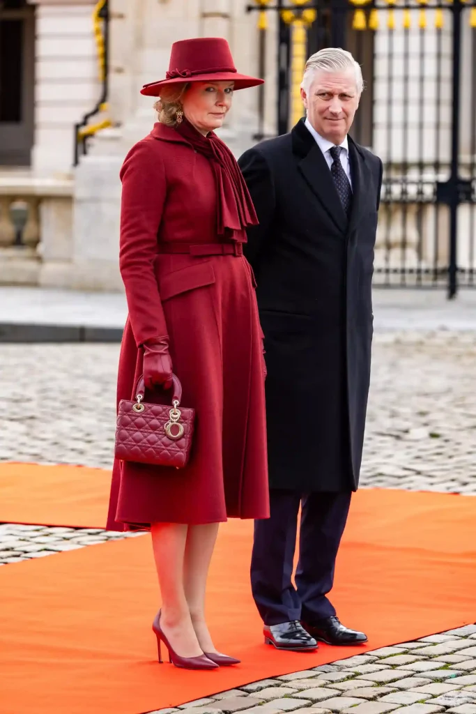 Queen Mathilde of Belgium in an elegant red coat and hat stands next to a man in a black coat on an orange carpet.